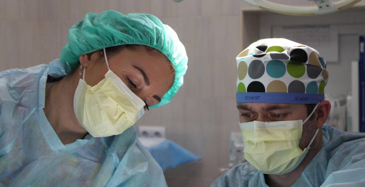 A team of focused medical professionals in a sterile operating room, with the lead surgeon making an incision while an assistant holds surgical instruments; monitors displaying vital signs are visible in the background