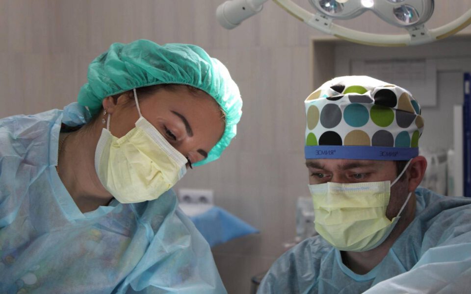 A team of focused medical professionals in a sterile operating room, with the lead surgeon making an incision while an assistant holds surgical instruments; monitors displaying vital signs are visible in the background